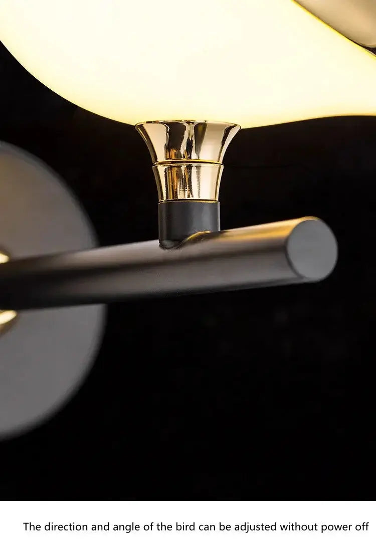 Close-up product detail of a modern wall lamp with a glowing warm white oval shade, a polished gold-toned neck, and a matte black horizontal arm against a dark background.
