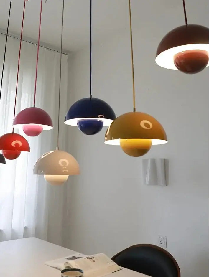 A set of colorful pendant dome lamps hanging over a dining table in a bright room. The lamps have glossy semi-spherical shades in red, burgundy, navy, mustard yellow, cream, and brown, with one lit up in warm white light.