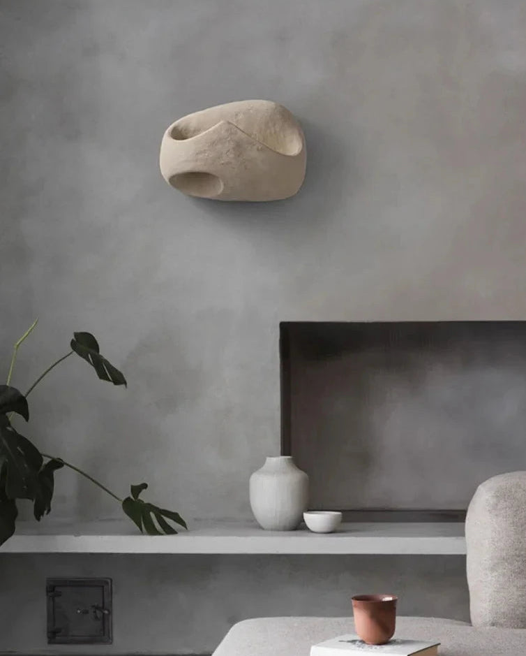 Hero A modern sculptural wall sconce in a light beige stone-like finish, mounted on a textured gray wall above a minimalist shelf with ceramic vases and a plant.