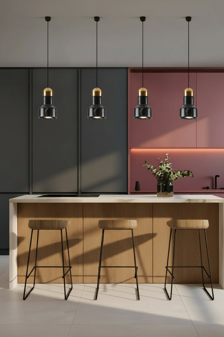 Modern kitchen interior with four black pendant lights featuring gold-toned caps hanging above a white island with three wooden bar stools. The kitchen cabinets are dark gray on the left and dusty rose on the right, with warm under-cabinet lighting.