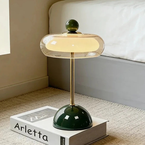 Hero A modern table lamp with a glowing warm yellow shade, a gold-finished stem, and a glossy dark green base, styled on top of a white book in a bedroom setting.