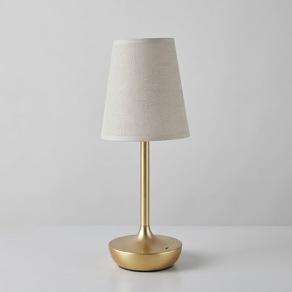 Hero A table lamp with a warm glowing fabric shade and a brushed gold metal base, shown on a tabletop beside a small glass vase with white flowers. The brand name KFV appears in the top left corner.