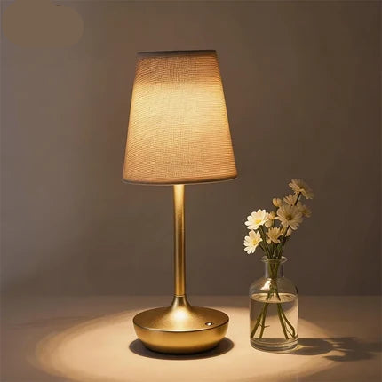A table lamp with a warm glowing fabric shade and a brushed gold metal base, shown on a tabletop beside a small glass vase with white flowers. The brand name KFV appears in the top left corner.