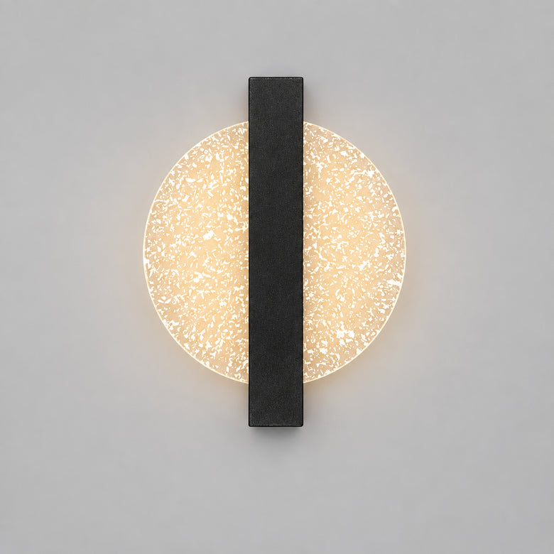 Hero A modern wall sconce with a circular textured glass shade and a central dark gray vertical bar, mounted on a beige wall above a shelf with decorative objects including a bust, candles, and a vase.