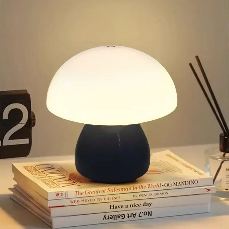 Hero A Black mushroom-shaped table lamp with a glowing white dome and a dark navy blue base, styled on top of stacked books on a tabletop with a clock and reed diffuser in the background.