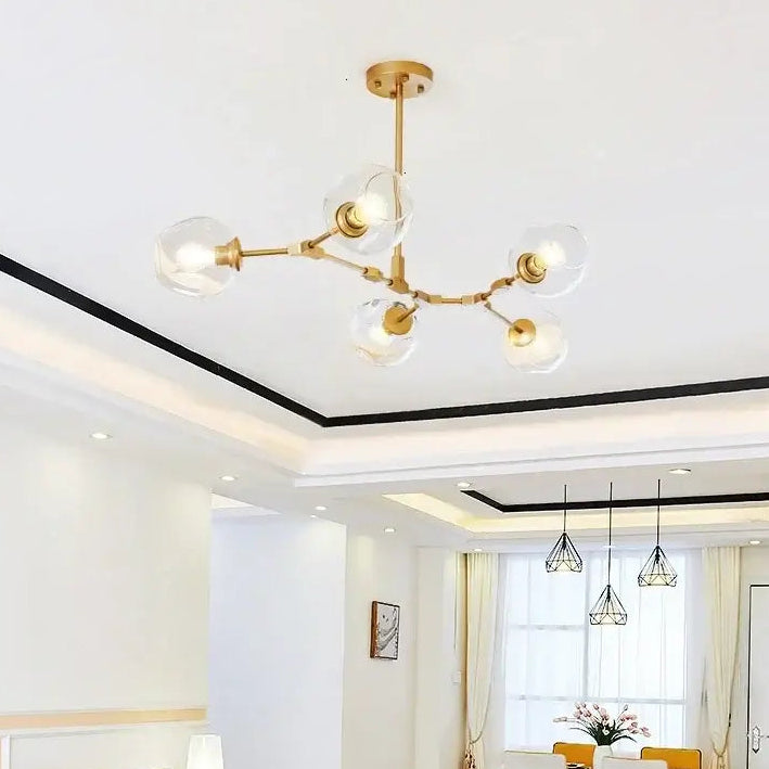 A modern brass multi-arm ceiling chandelier with clear glass globe shades hanging in a bright, contemporary living room with a sofa, coffee table, and dining area in the background.