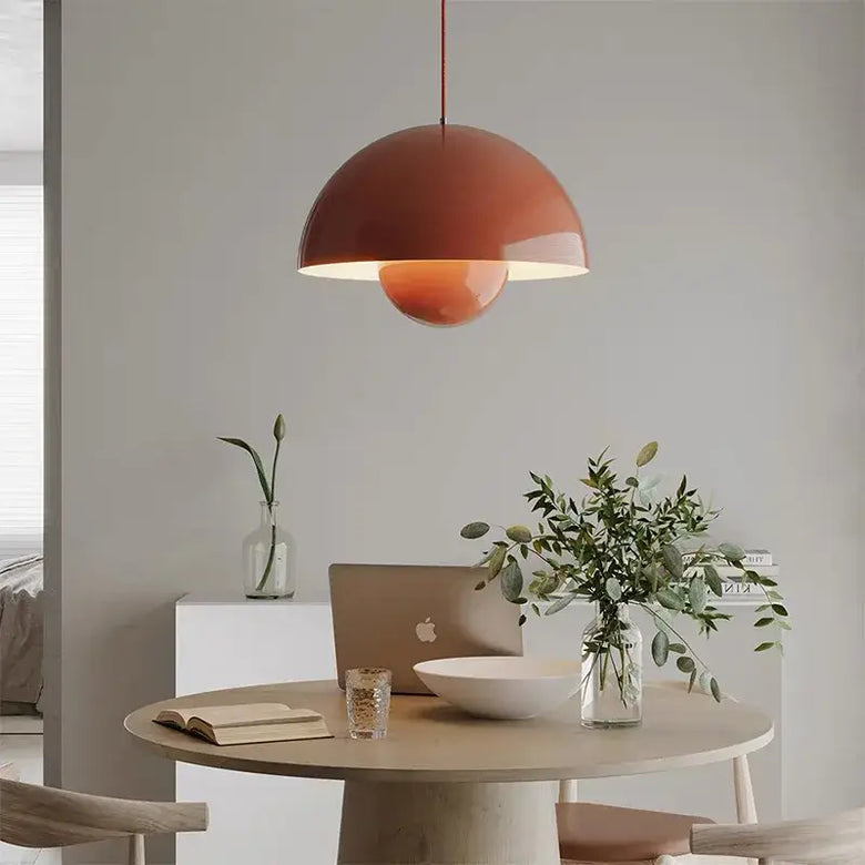 A modern terracotta-colored dome pendant light hanging over a round wooden dining table in a minimalist, neutral-toned room. The table is styled with a laptop, a bowl, a glass, and a vase of greenery.