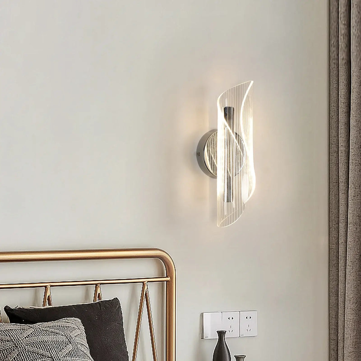 Contemporary chrome ribbon LED wall light with a clear acrylic shade, lit above a bed