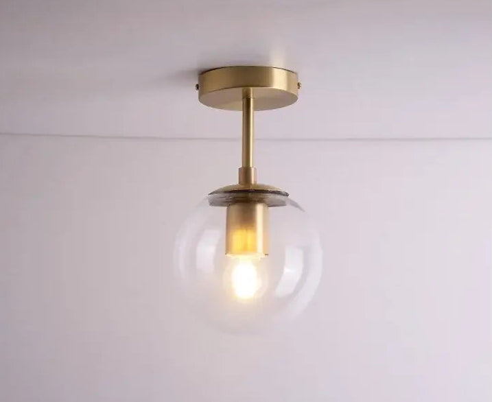 A modern semi-flush mount ceiling light fixture with a brushed brass/gold metal canopy and stem, featuring a clear glass globe shade that illuminates a warm light bulb.