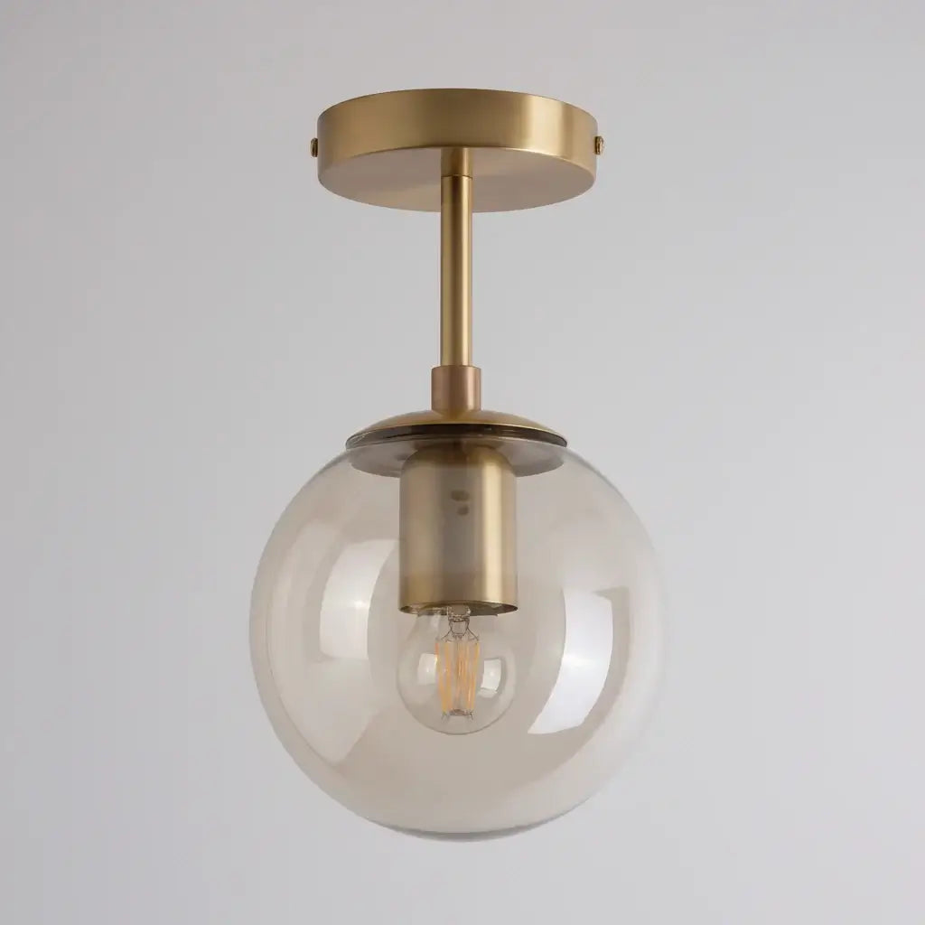 Hero A modern semi-flush mount ceiling light fixture featuring a large, clear glass globe shade and a brushed brass metal canopy and stem, with the warm glow of an exposed filament light bulb visible inside.