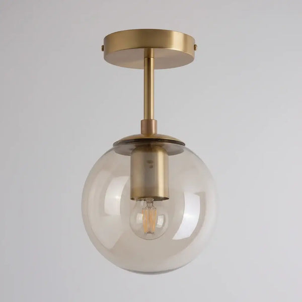 Hero A modern semi-flush mount ceiling light fixture featuring a large, clear glass globe shade and a brushed brass metal canopy and stem, with the warm glow of an exposed filament light bulb visible inside.