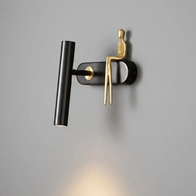 Hero A modern wall-mounted light fixture with a matte black cylindrical lamp head and a sculptural gold figure sitting on a black base, shown against a dark gray wall with warm light shining downward.