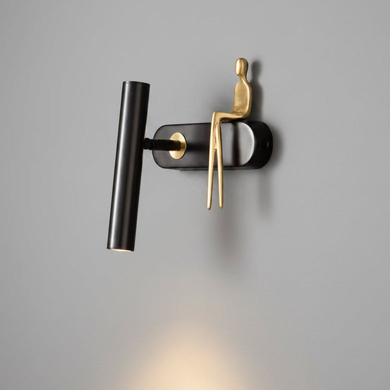 Hero A modern wall-mounted light fixture with a matte black cylindrical lamp head and a sculptural gold figure sitting on a black base, shown against a dark gray wall with warm light shining downward.
