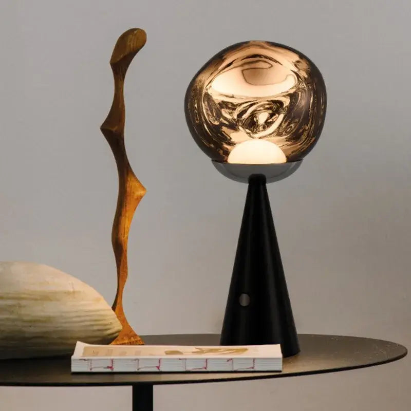 A modern table lamp with a glossy, reflective metallic gold globe shade on a black cone base, styled on a dark round table next to a wooden sculpture and a book.