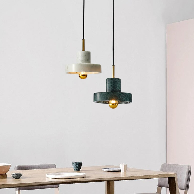 Two modern pendant lights hanging above a wooden dining table in a minimalist room. One light has a white marble-look top and shade with a gold center, and the other has a dark green marble-look top and shade with a gold center.