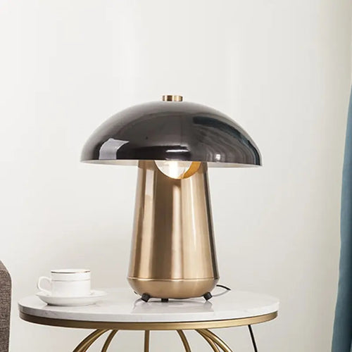 A modern mushroom-shaped table lamp with a glossy black dome shade and a brushed gold metallic base, styled on a white marble-topped side table next to a white cup and saucer.