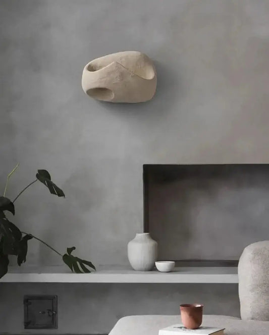 Hero A modern sculptural wall sconce in a light beige stone-like finish, mounted on a textured gray wall above a minimalist shelf with ceramic vases and a plant.