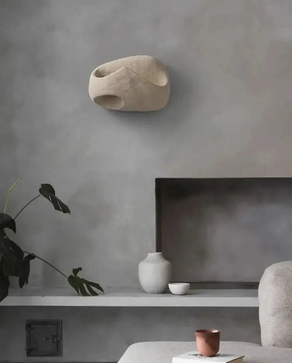 Hero A modern sculptural wall sconce in a light beige stone-like finish, mounted on a textured gray wall above a minimalist shelf with ceramic vases and a plant.