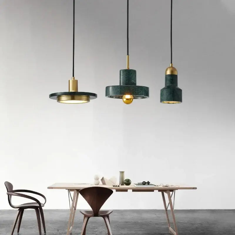 Three modern pendant lights hang above a dining table in a minimalist room. The lights have dark green cylindrical shades with gold-toned metal accents and black cords, set against a clean white wall.