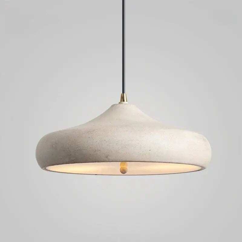 A modern pendant light with a wide, shallow dome-shaped shade made of textured concrete in a warm off-white color. The light is illuminated from within, casting a warm glow, and hangs from a thin black cord with a brass connector against a neutral gray background.