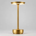Gold Art Deco cordless disc table lamp lit on a wooden surface with books and a plant