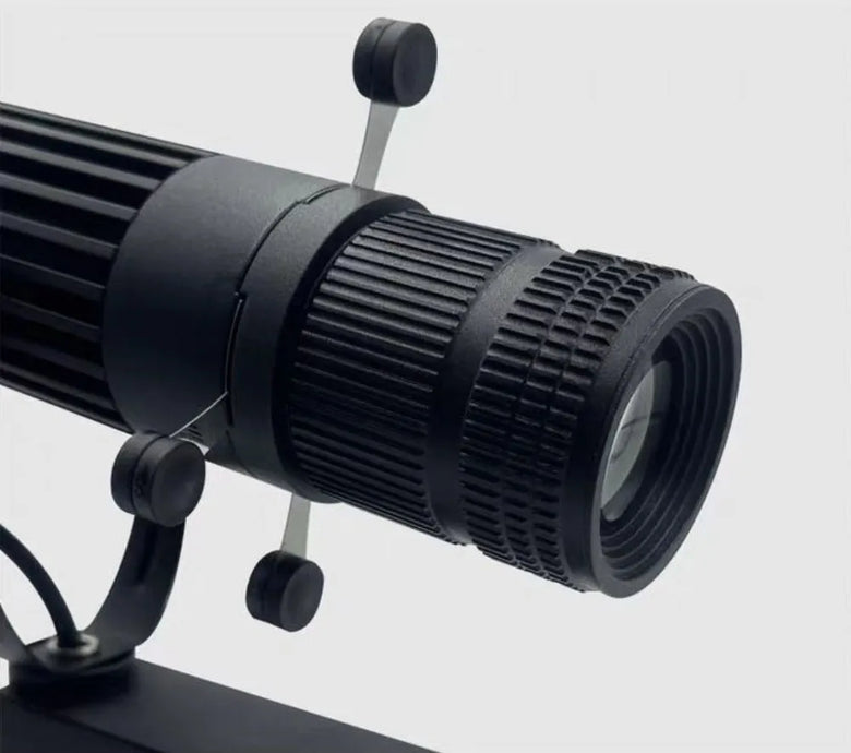 Close-up of a black optical device, likely a microscope or telescope eyepiece and objective lens assembly, mounted on a stand against a light gray background.