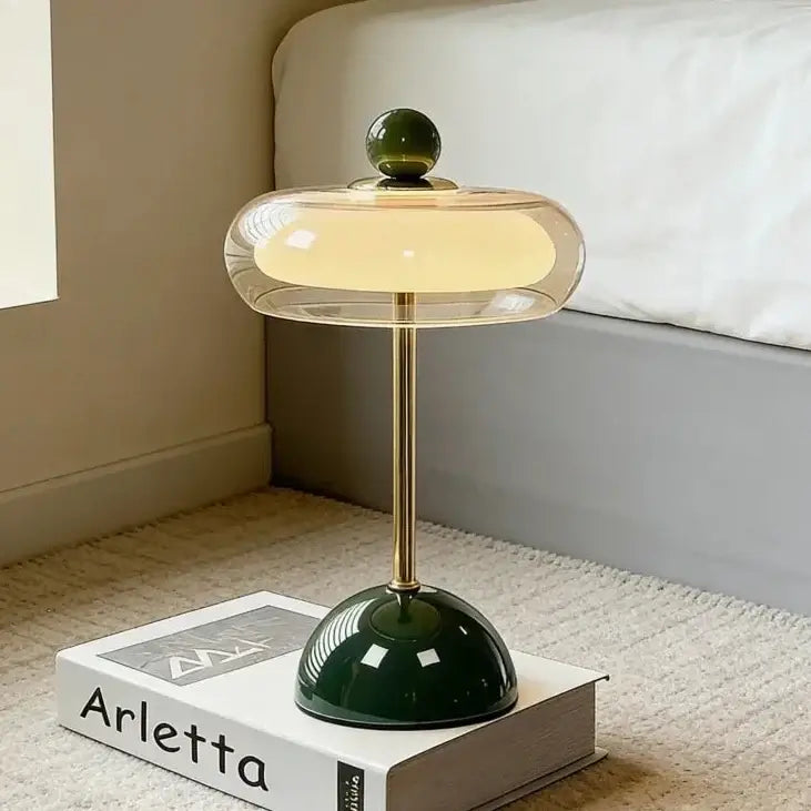 Hero A modern table lamp with a glowing warm yellow shade, a gold-finished stem, and a glossy dark green base, styled on top of a white book in a bedroom setting.