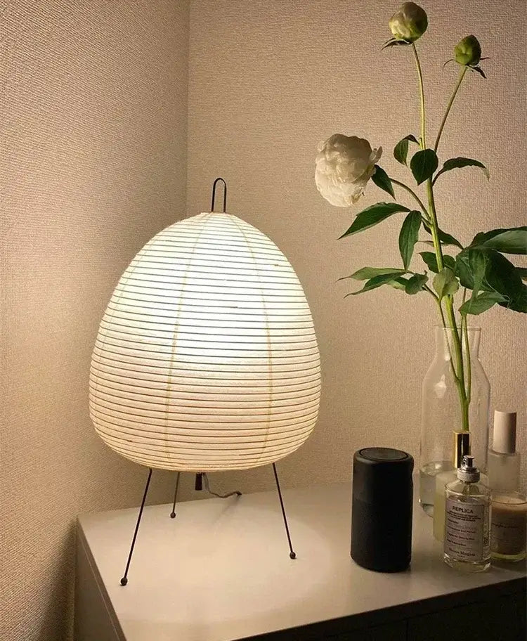 Japanese Rice Paper Lamp. White woven table lamp on a surface with a vase of flowers and other items in the background.