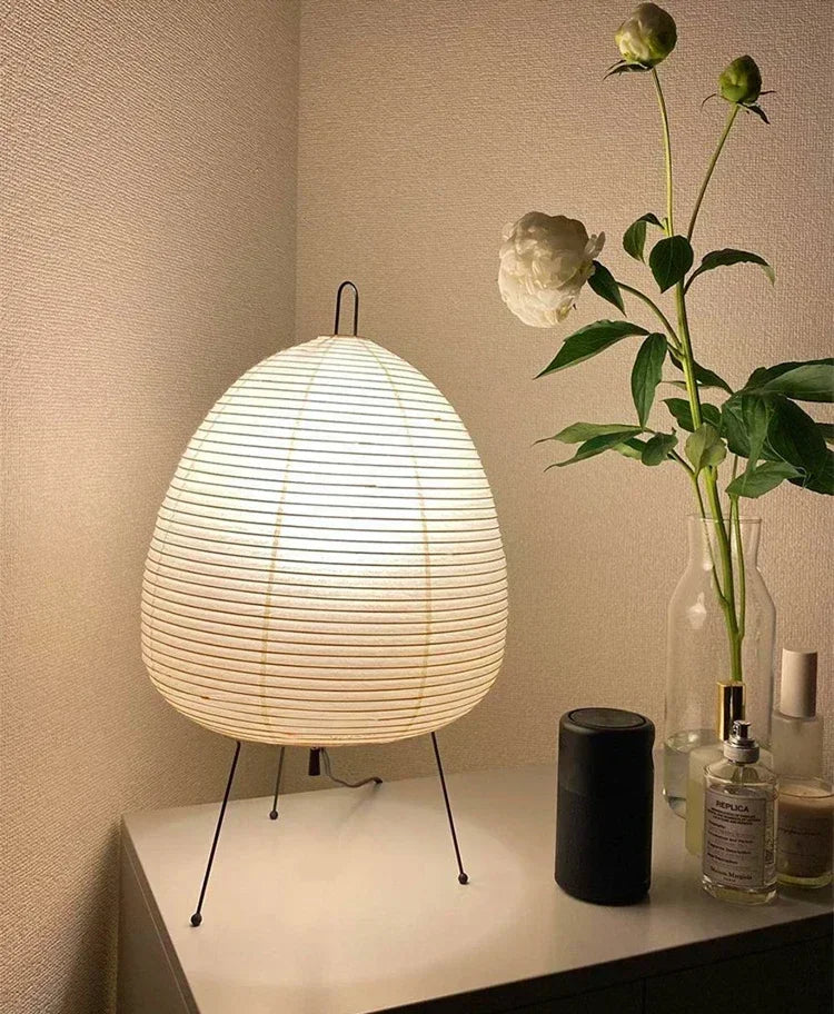 Japanese Rice Paper Lamp. White woven table lamp on a surface with a vase of flowers and other items in the background.