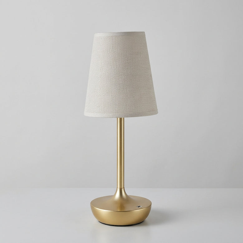 Hero A table lamp with a warm glowing fabric shade and a brushed gold metal base, shown on a tabletop beside a small glass vase with white flowers. The brand name KFV appears in the top left corner.