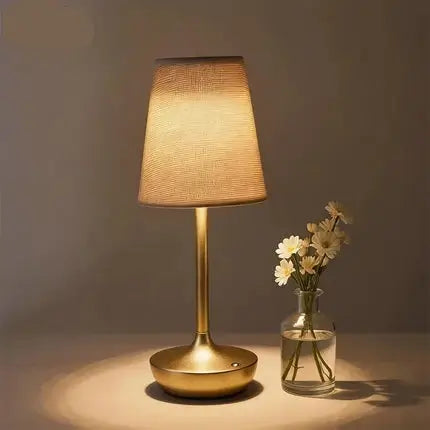 A table lamp with a warm glowing fabric shade and a brushed gold metal base, shown on a tabletop beside a small glass vase with white flowers. The brand name KFV appears in the top left corner.