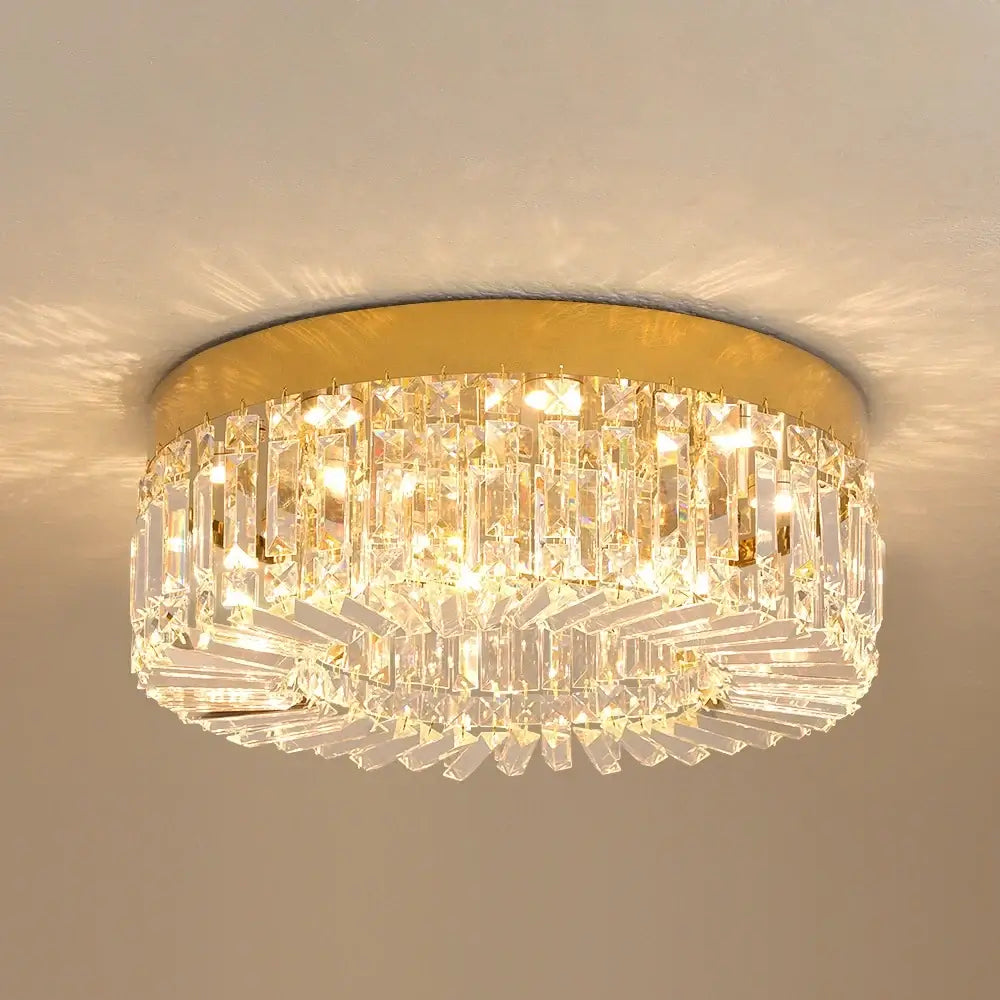 Hero A flush-mount ceiling light fixture with a gold circular metal frame and multiple tiers of clear crystal prisms, illuminated against a neutral beige wall.