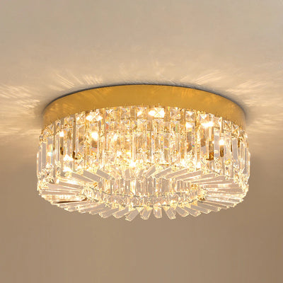 Hero A flush-mount ceiling light fixture with a gold circular metal frame and multiple tiers of clear crystal prisms, illuminated against a neutral beige wall.