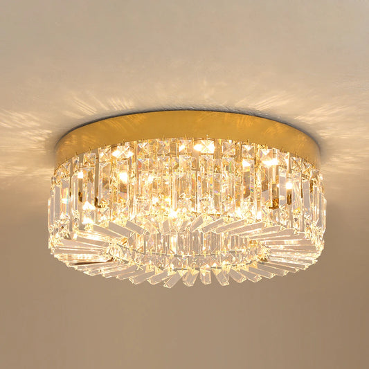 Hero A flush-mount ceiling light fixture with a gold circular metal frame and multiple tiers of clear crystal prisms, illuminated against a neutral beige wall.