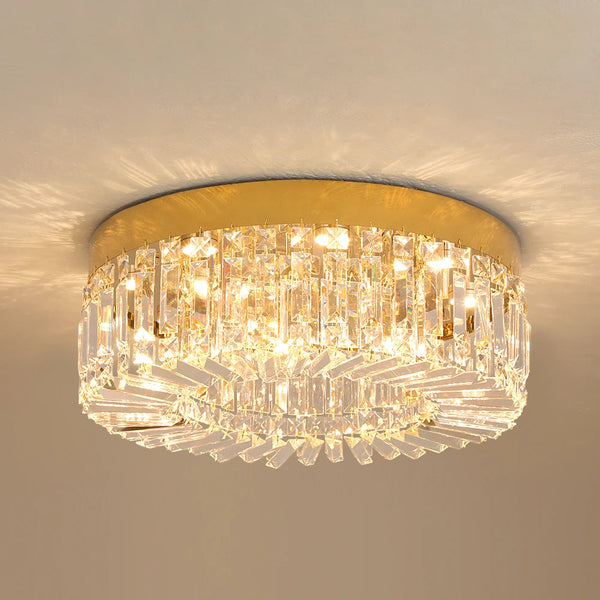 Hero A flush-mount ceiling light fixture with a gold circular metal frame and multiple tiers of clear crystal prisms, illuminated against a neutral beige wall.