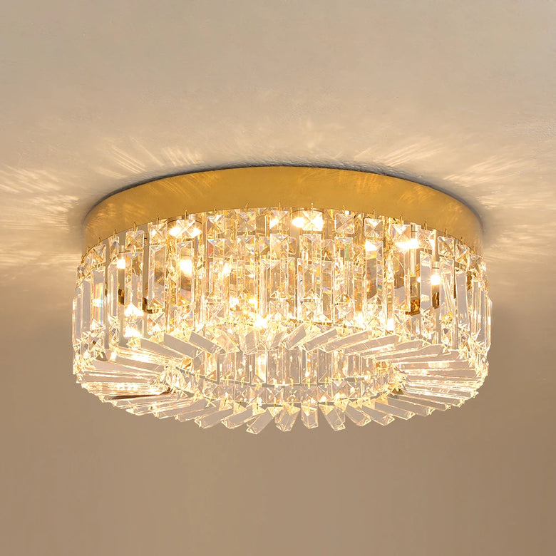 Hero A flush-mount ceiling light fixture with a gold circular metal frame and multiple tiers of clear crystal prisms, illuminated against a neutral beige wall.