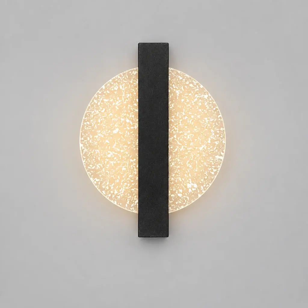 Hero A modern wall sconce with a circular textured glass shade and a central dark gray vertical bar, mounted on a beige wall above a shelf with decorative objects including a bust, candles, and a vase.