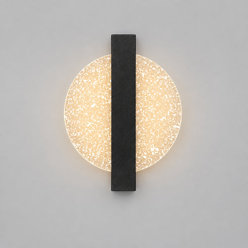 Hero A modern wall sconce with a circular textured glass shade and a central dark gray vertical bar, mounted on a beige wall above a shelf with decorative objects including a bust, candles, and a vase.