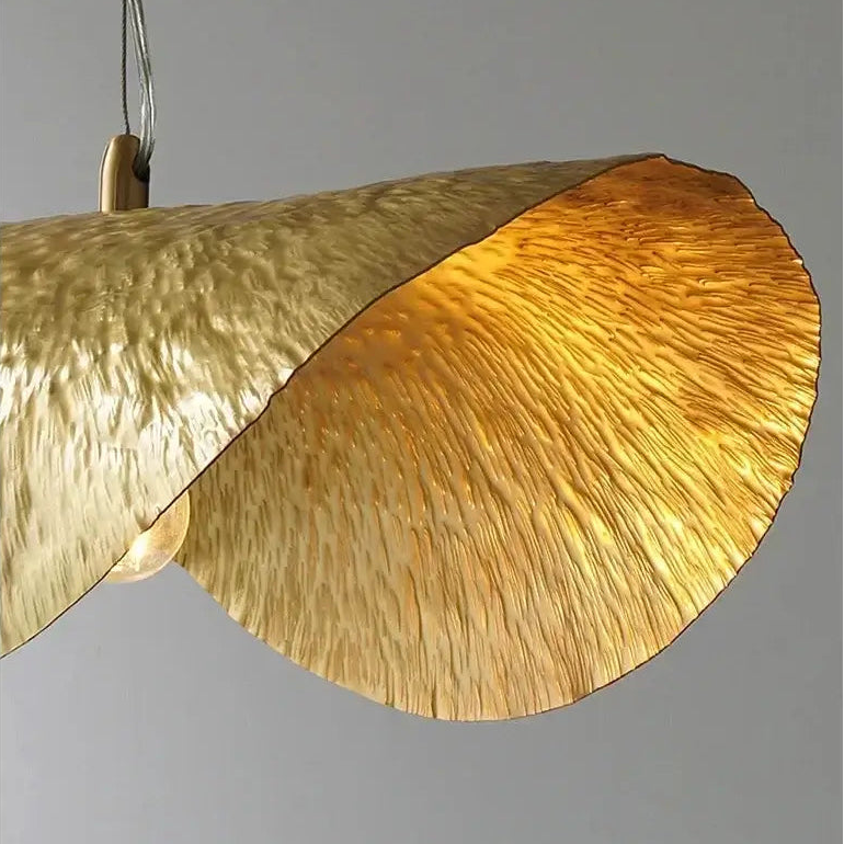 hero A close-up of a modern pendant light with a large, textured metallic gold shade that has a wavy, layered design, shown against a solid gray background.