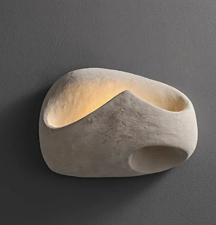 An organic-shaped wall sconce made of textured stone or concrete, featuring a warm internal light glowing from an open cavity, mounted on a dark gray wall.