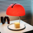 80s USB desk lamp in various colours with a red dome shade and chrome base next to a candle on a white table.