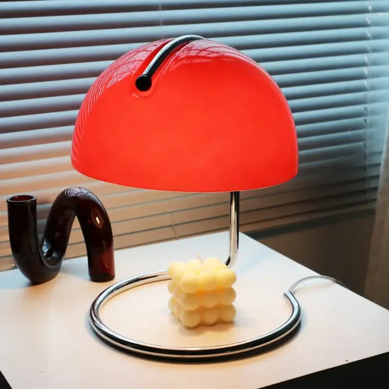 80s USB desk lamp in various colours with a red dome shade and chrome base next to a candle on a white table.