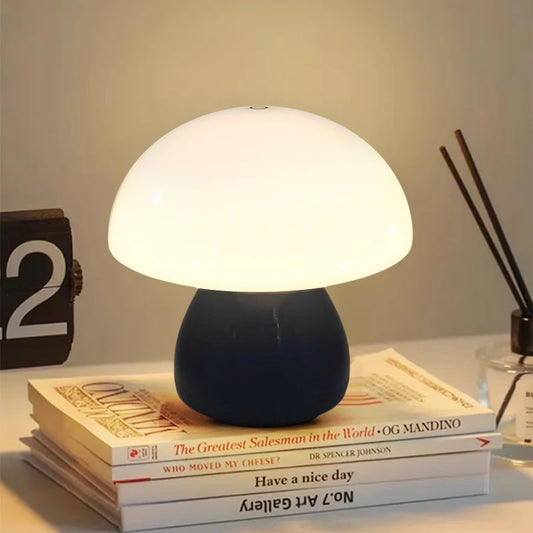 Hero A Black mushroom-shaped table lamp with a glowing white dome and a dark navy blue base, styled on top of stacked books on a tabletop with a clock and reed diffuser in the background.