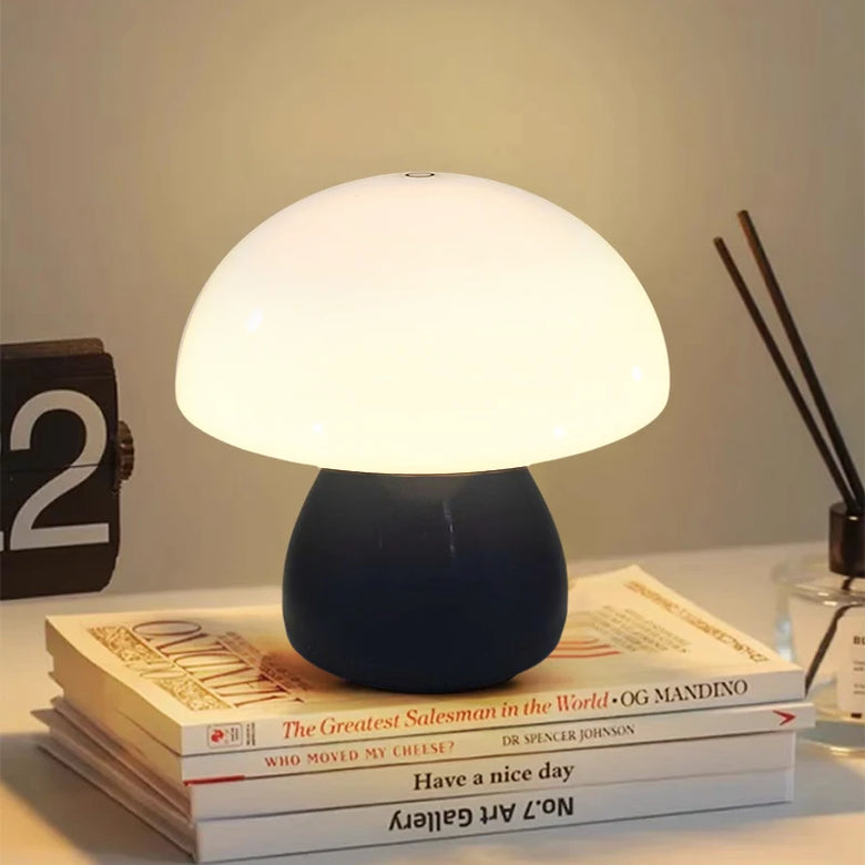 Hero A Black mushroom-shaped table lamp with a glowing white dome and a dark navy blue base, styled on top of stacked books on a tabletop with a clock and reed diffuser in the background.