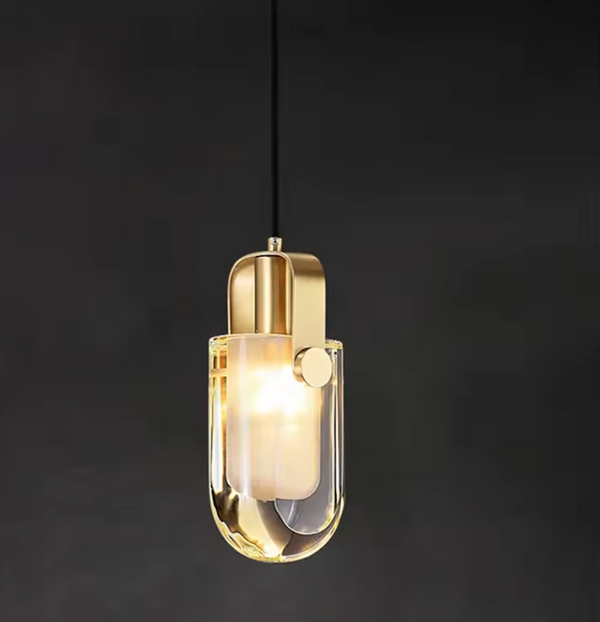 Hero A modern pendant light with a polished gold-finished metal body and a clear glass cylindrical shade, glowing with a warm light against a dark background.