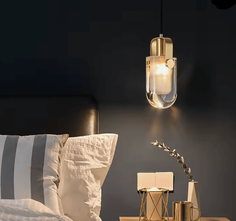 A modern pendant light with a gold-finished metal body and clear glass shade, glowing warmly in a bedroom setting above a bed with striped pillows.