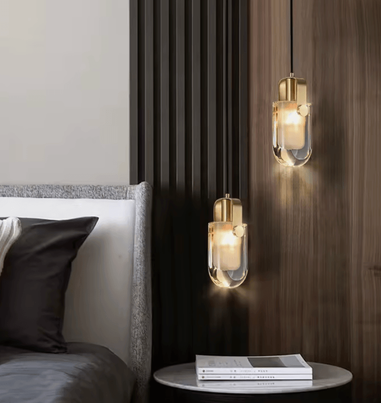 Two modern pendant light fixtures with clear glass cylindrical shades and brushed gold caps, hanging in a bedroom setting next to a bed and above a small side table.