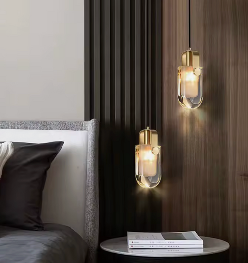 Two modern pendant light fixtures with clear glass cylindrical shades and brushed gold caps, hanging in a bedroom setting next to a bed and above a small side table.