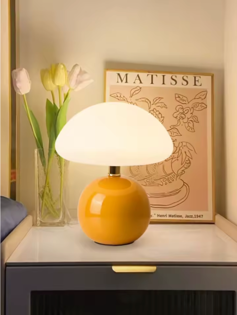 hero - A yellow mushroom-shaped table lamp with a glossy yellow-orange base and a glowing white dome shade, styled on a bedside table next to a vase of tulips and a framed Matisse print.