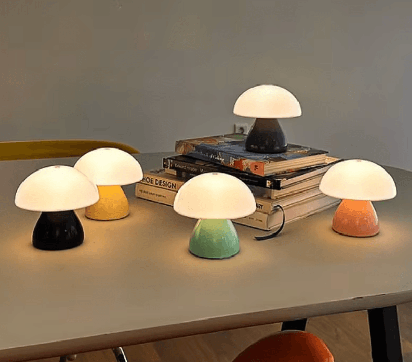 Five mushroom-shaped table lamps in different colors (black, yellow, dark gray, mint green, and peach) arranged on a tabletop with books in the background.